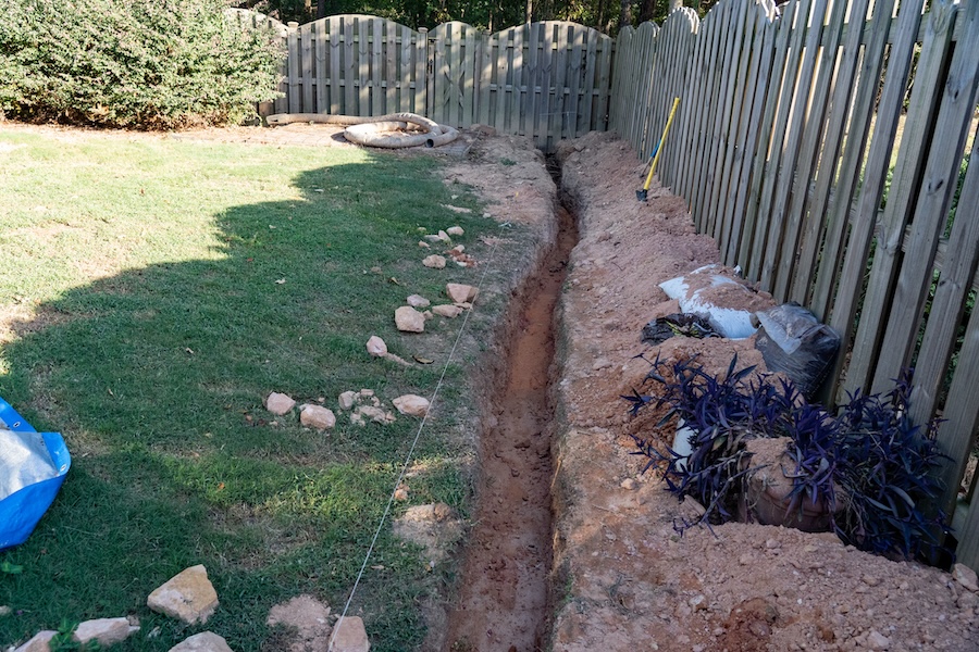 Trench excavated through a backyard of red clay for the DIY installation of a french drain.